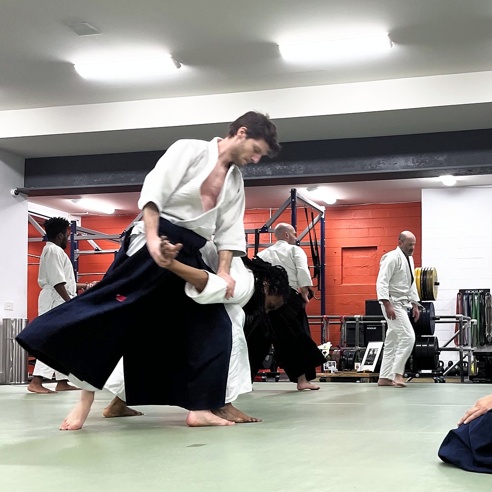 A student bending his partner forward with an arm lock while the instructor kneels and watches
